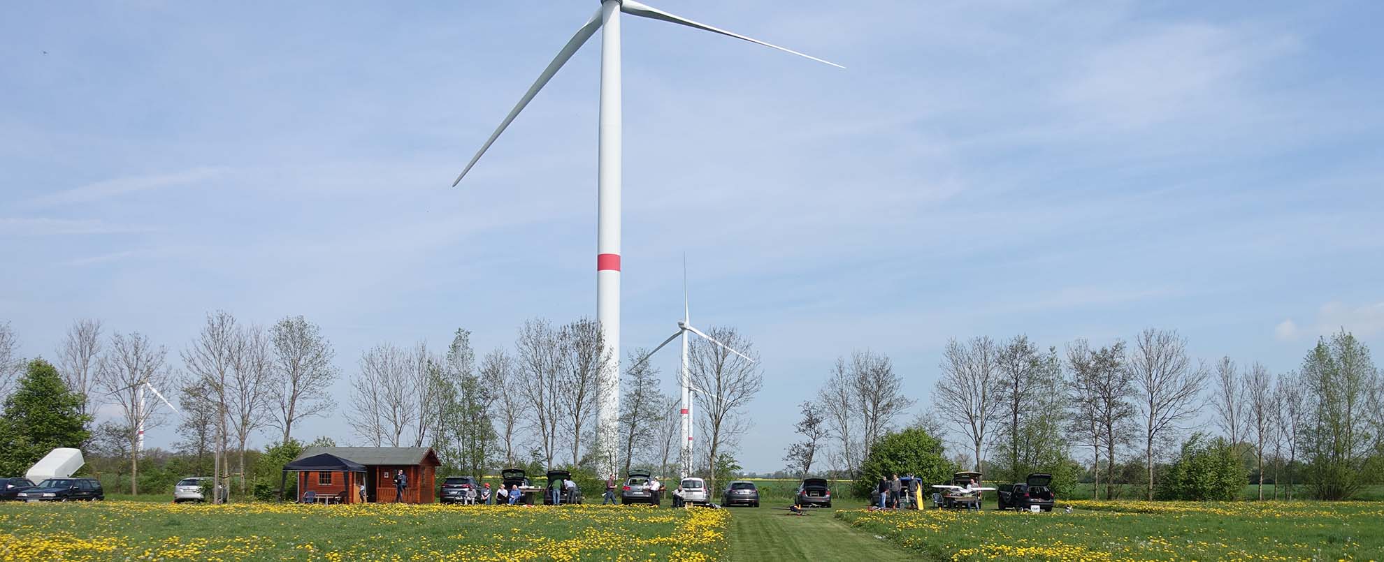 Landschaftsbild einer Wiese mit einem roten Gebäude, einigen Autos und einer Windmühle im Hintergrund